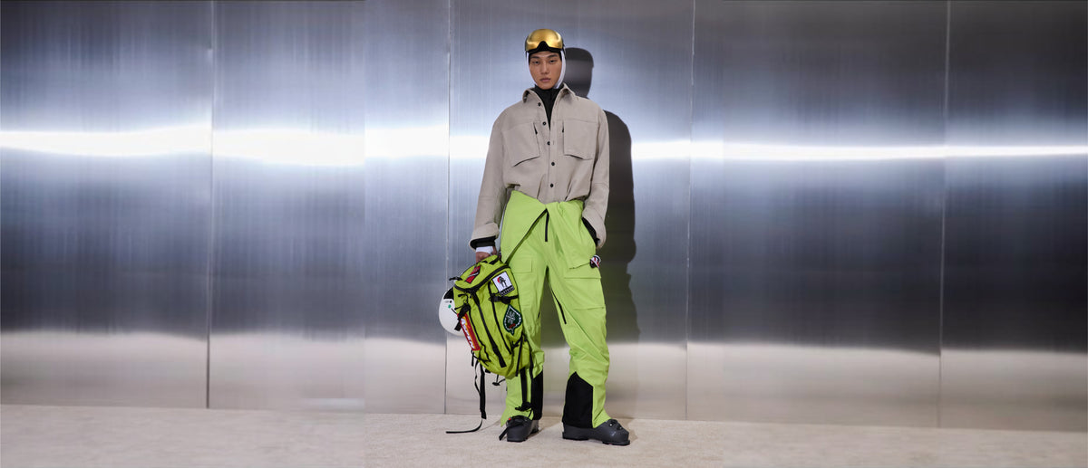 Person wearing a beige jacket and anti-freeze color Hayden 3L bibs standing in front of a metallic wall holding a backpack and helmet