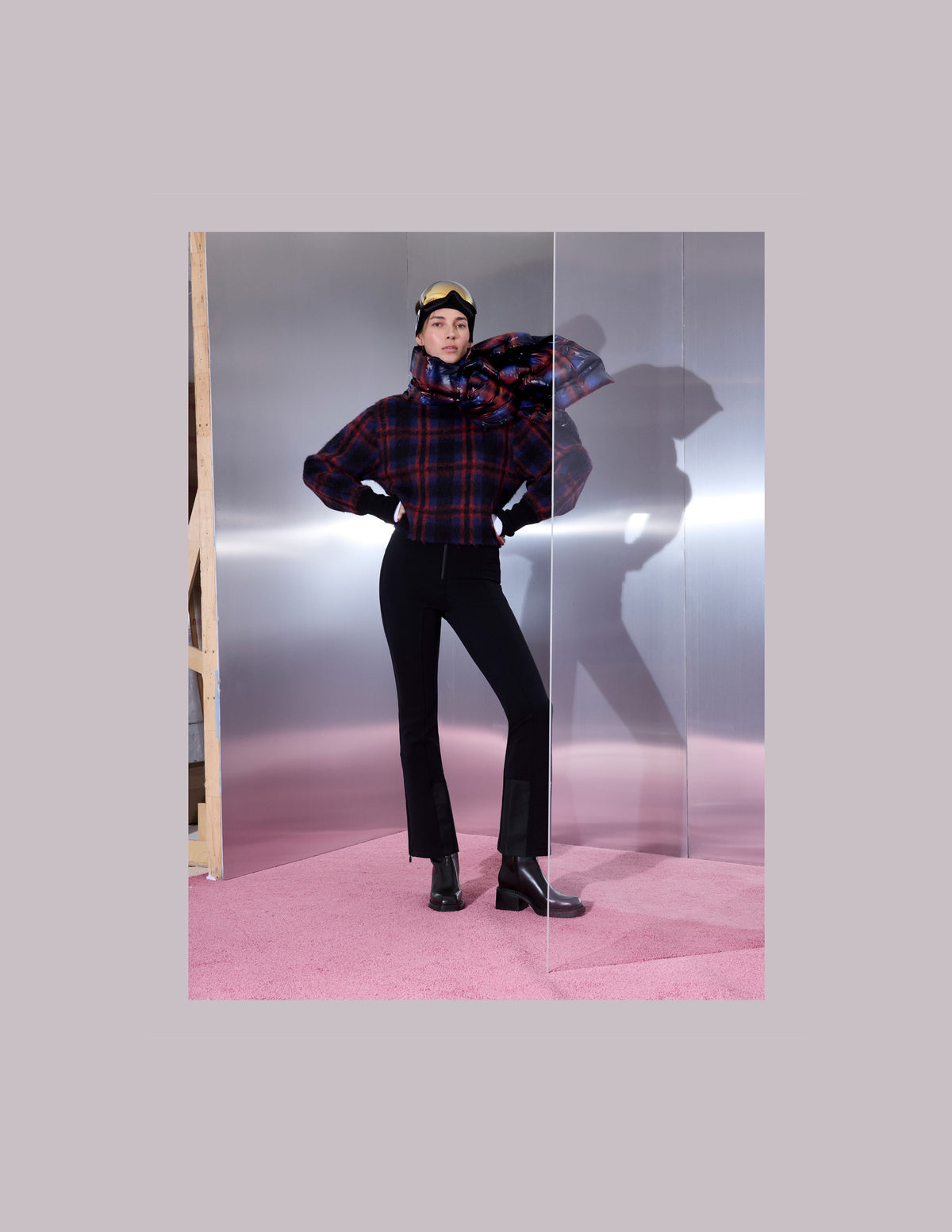 A model stands in a studio setting wearing a dark red and navy plaid jacket with a matching scarf, black pants, and black boots.