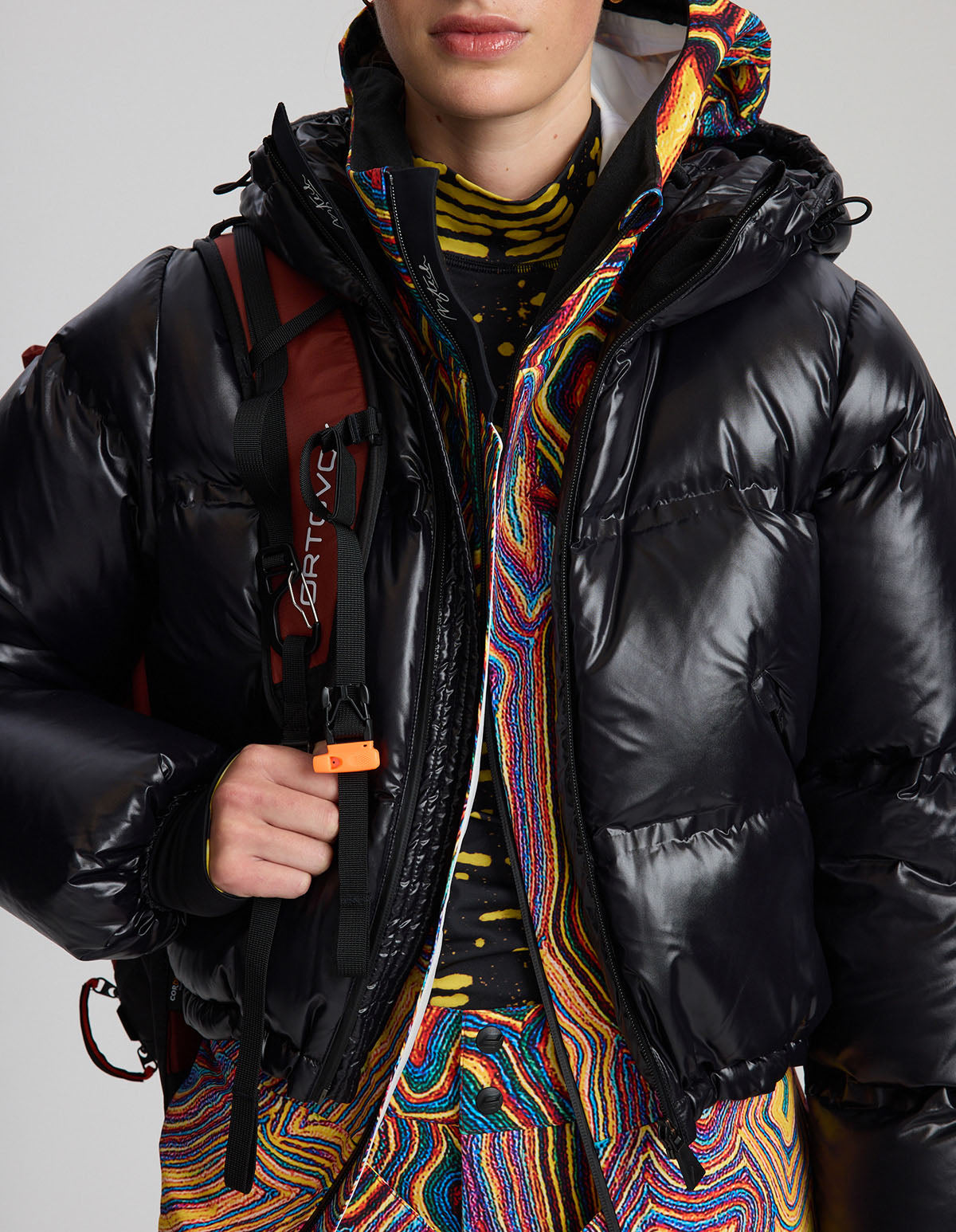 Woman wearing a black puffer jacket with colorful patterns and a backpack on a gray background