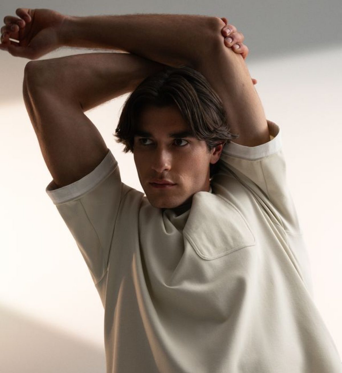 A man with dark hair poses with his arms raised over his head, wearing a light beige short-sleeve top with a large chest pocket, set against a minimalist off-white background.