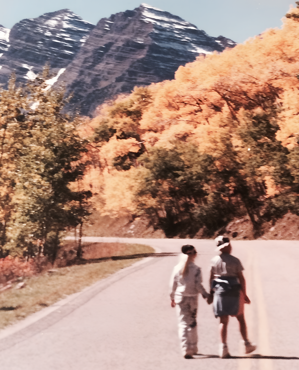 Two people walking on a road with mountains and autumn trees in the background
