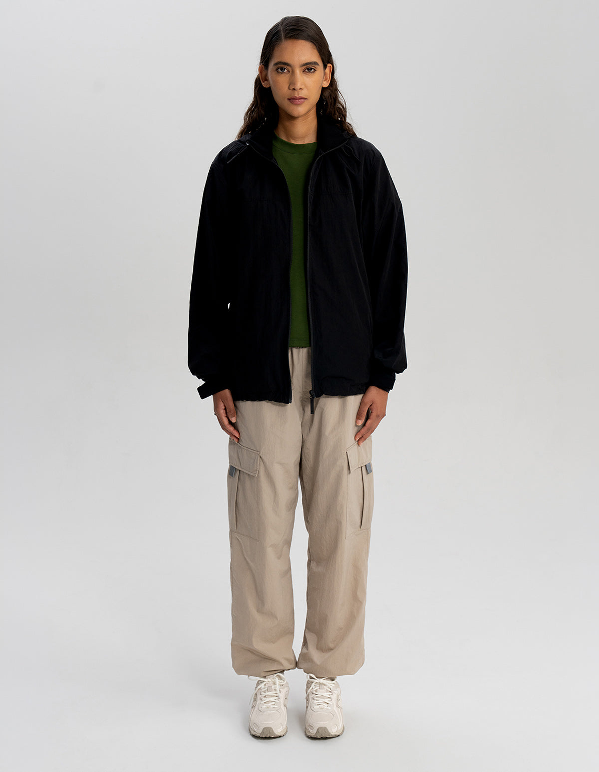 A model stands against a light gray studio background wearing a black zip-up jacket over a dark green top, beige cargo pants, and white sneakers. 