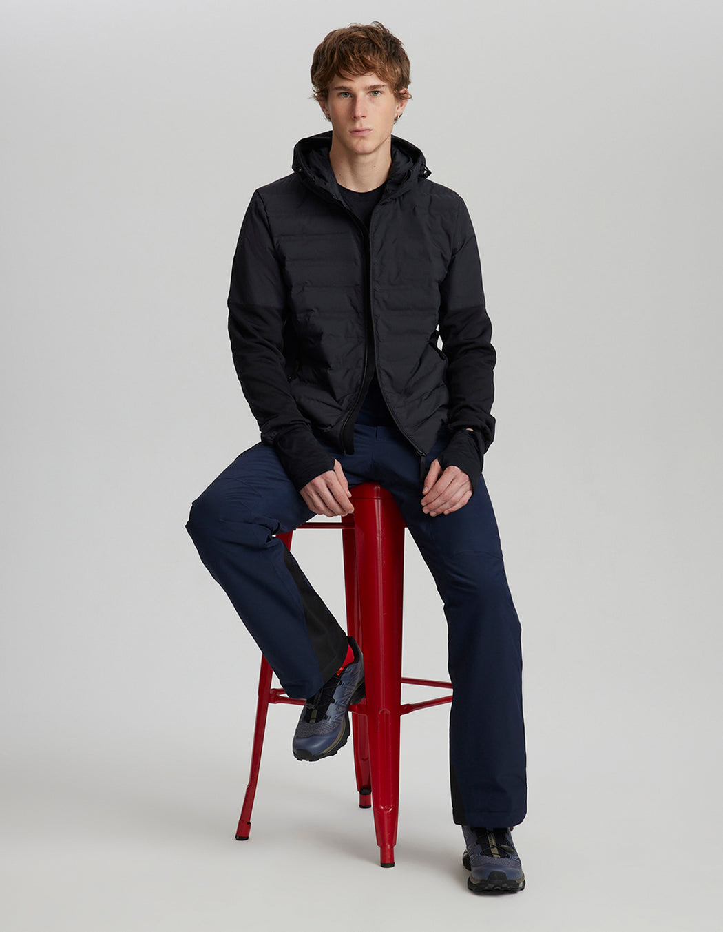 Man sitting on a red stool wearing a black jacket and navy pants against a white background
