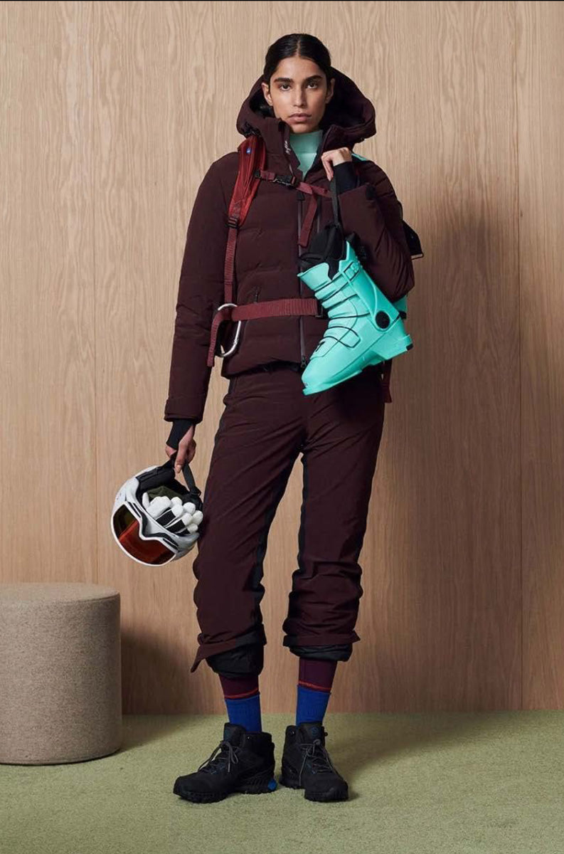 A model stands against a light wood-paneled wall wearing a dark brown ski jacket and matching pants, with a turquoise ski boot resting on her shoulder and a white helmet in her hand.