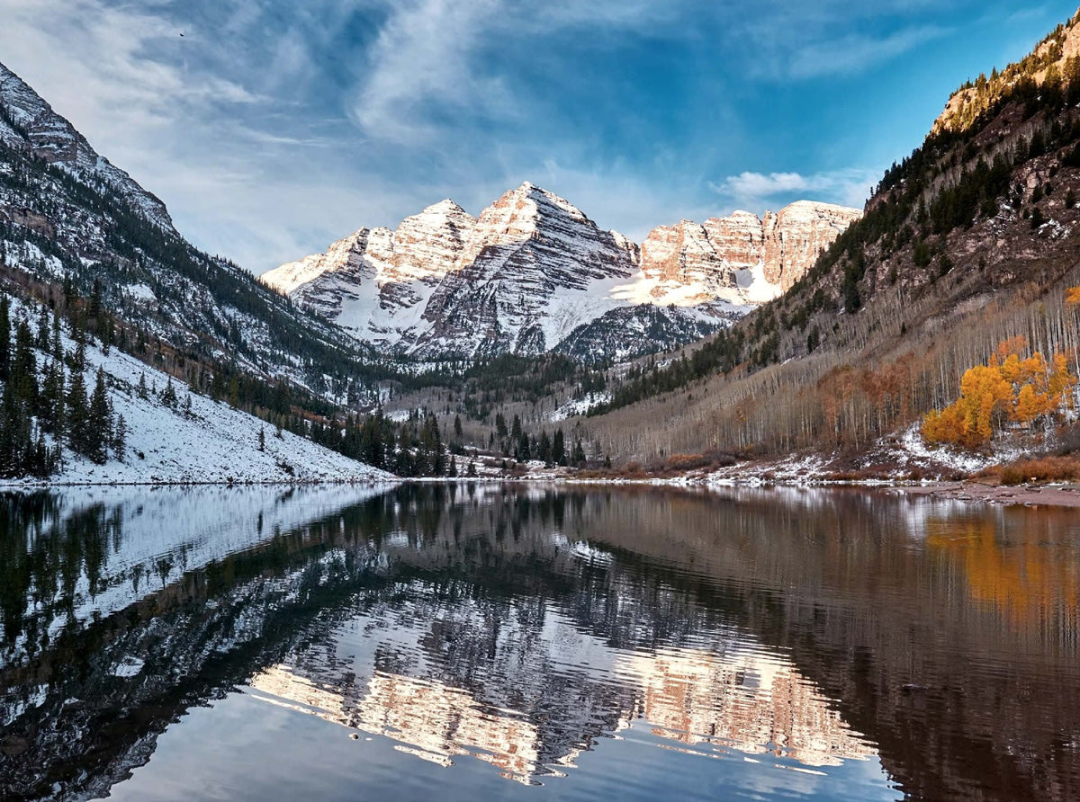 A scenic mountain landscape with snow-covered peaks reflected in a calm lake, surrounded by evergreen and autumn-colored trees under a blue sky with light clouds.