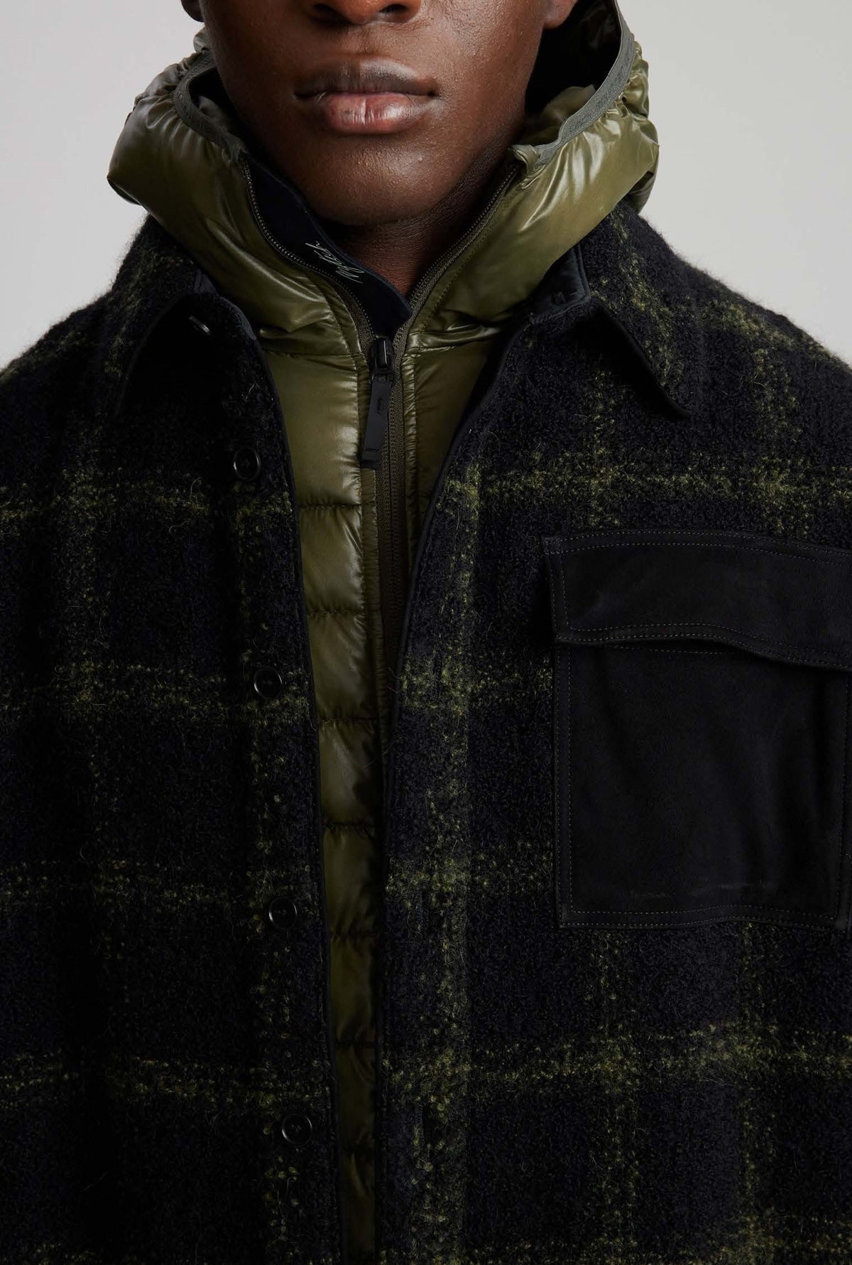 Close-up of a person wearing a dark plaid outer layer over an olive green puffer jacket, with the lower face and upper torso visible against a plain light background.