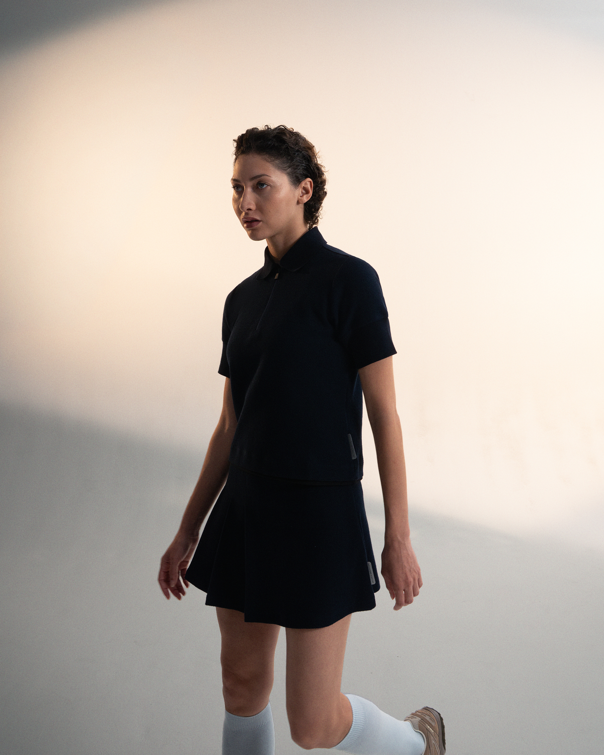 A woman modeling a dark navy athletic set consisting of a short-sleeve collared top and a matching mini skirt, paired with white knee-high socks and brown sneakers, set against a minimalist beige studio background.