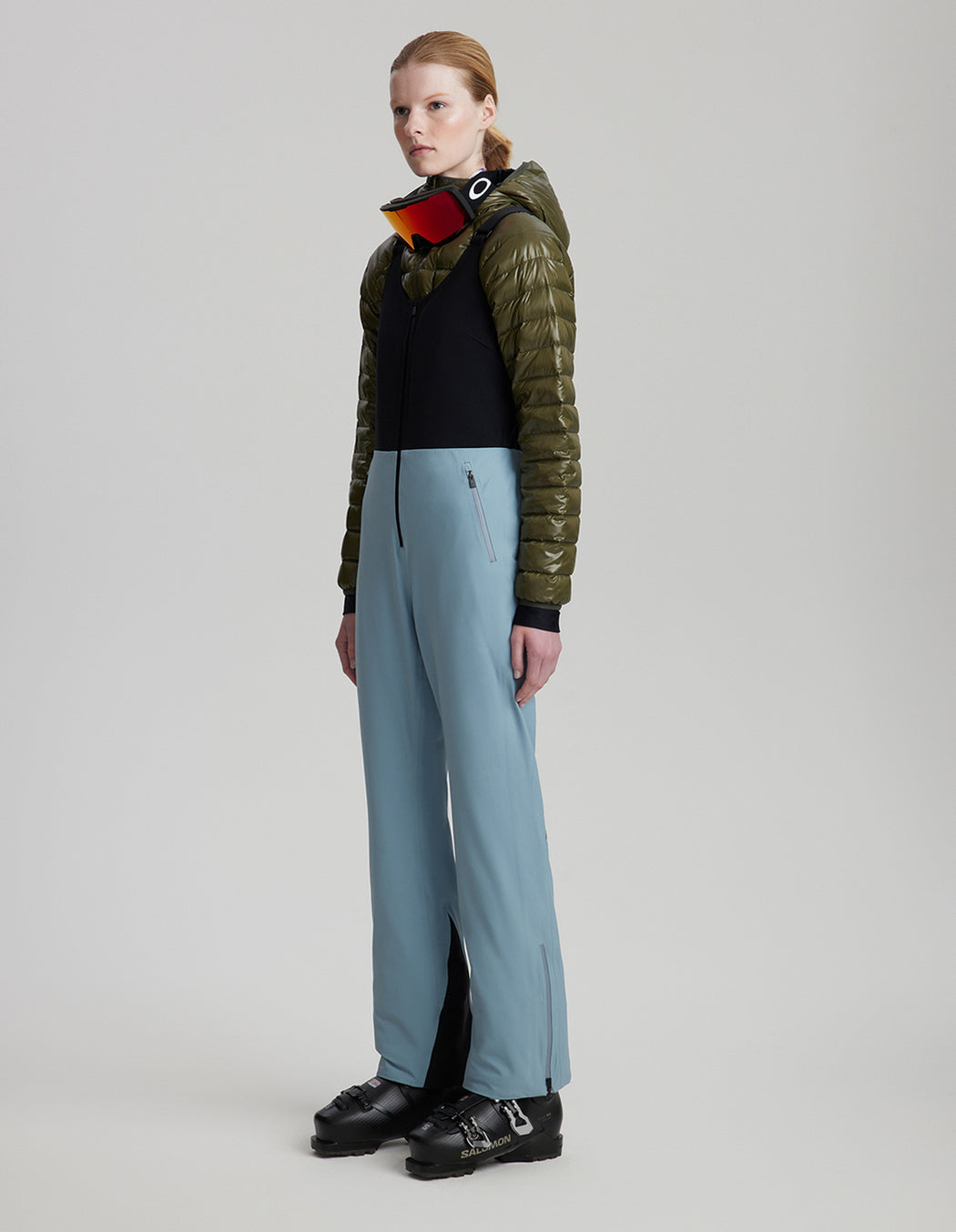 Person wearing a green jacket, black shirt, light blue pants, and black shoes on a gray background