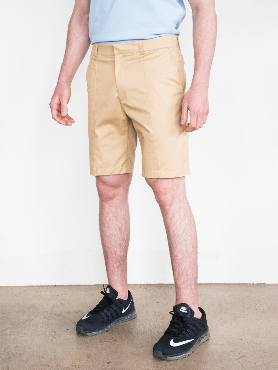 Khaki tailored shorts with flat front and side pockets worn with black sneakers and light blue shirt.
