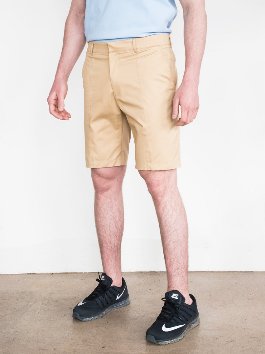 Khaki tailored shorts with flat front and side pockets worn with black sneakers and light blue shirt.